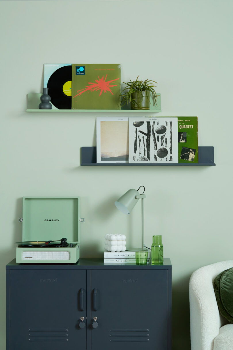 A modern living room scene with a green record player on a dark green cabinet, shelves displaying vinyl records and plants, and various small decor items