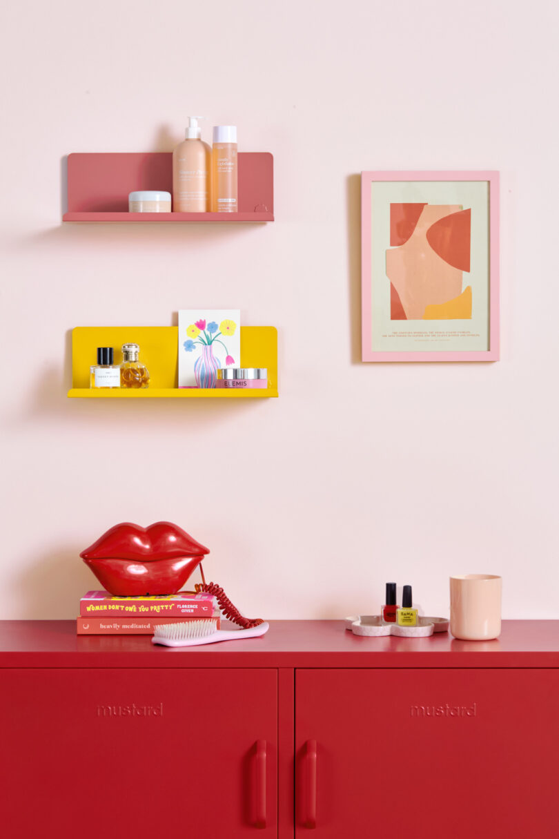 A red cabinet with various beauty products and a red lips phone on top. Two colorful wall shelves above hold more products, a card, and a framed abstract art piece. A mug is also on the cabinet