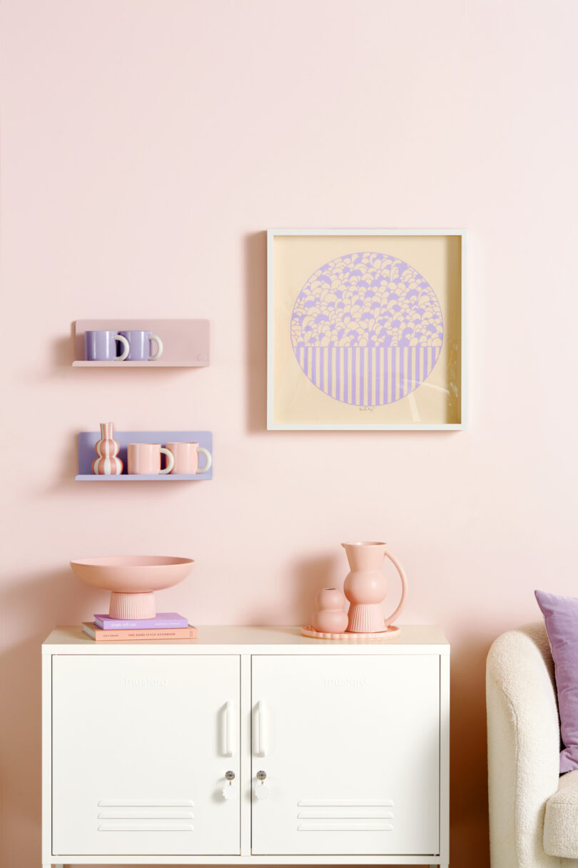 A pale pink wall with a framed geometric art piece, two shelves holding pastel mugs and vases, and a white cabinet with pink decor items underneath. A portion of a cream armchair is visible on the right