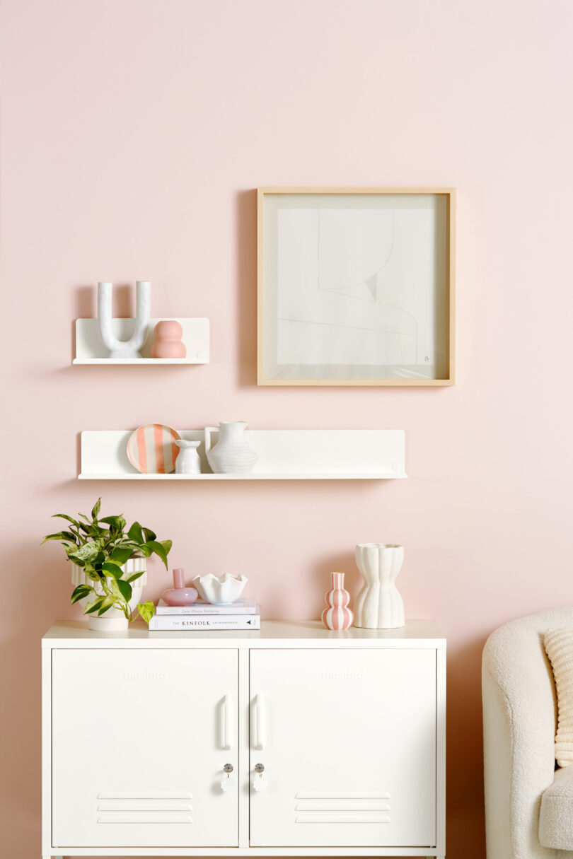 Pastel pink wall with minimalistic decor, including two floating shelves holding various vases, a framed abstract artwork, and a white cabinet with books, decorative items, and a plant