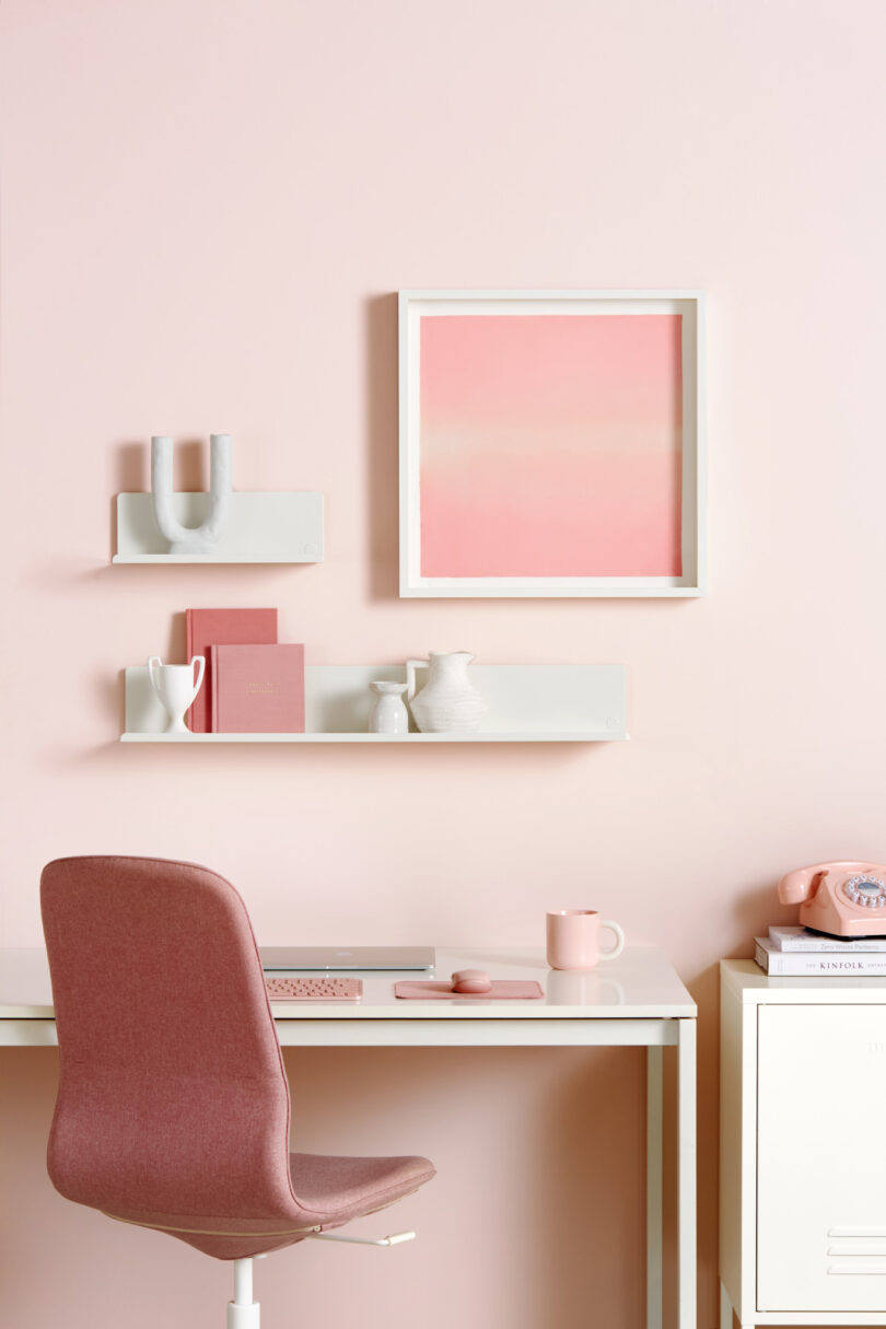 A pink-themed workspace with a desk, chair, books, a keyboard, a mouse, a mug, and decorative items on shelves against a pink wall with a framed abstract artwork