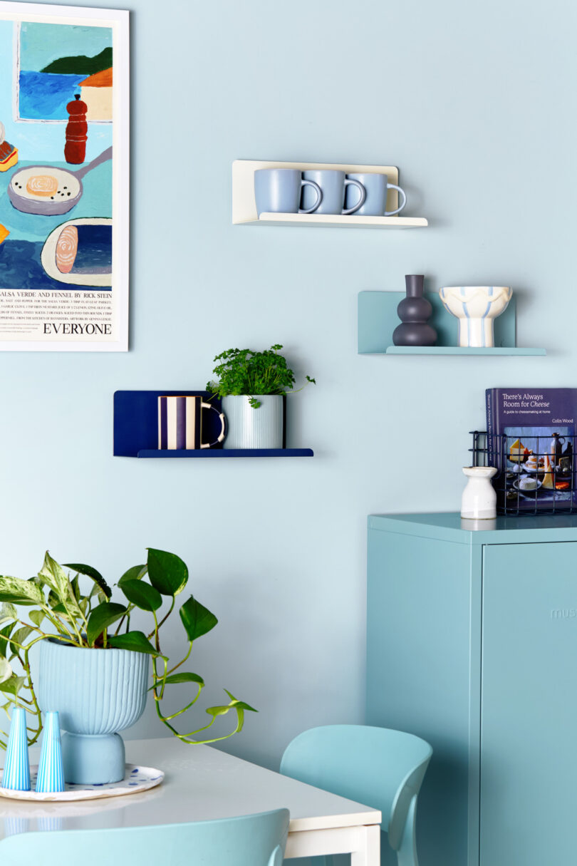 A kitchen scene featuring blue-toned walls, floating shelves with mugs and decor, a small plant, a potted plant on a white table, a teal chair, and a colorful poster on the wall