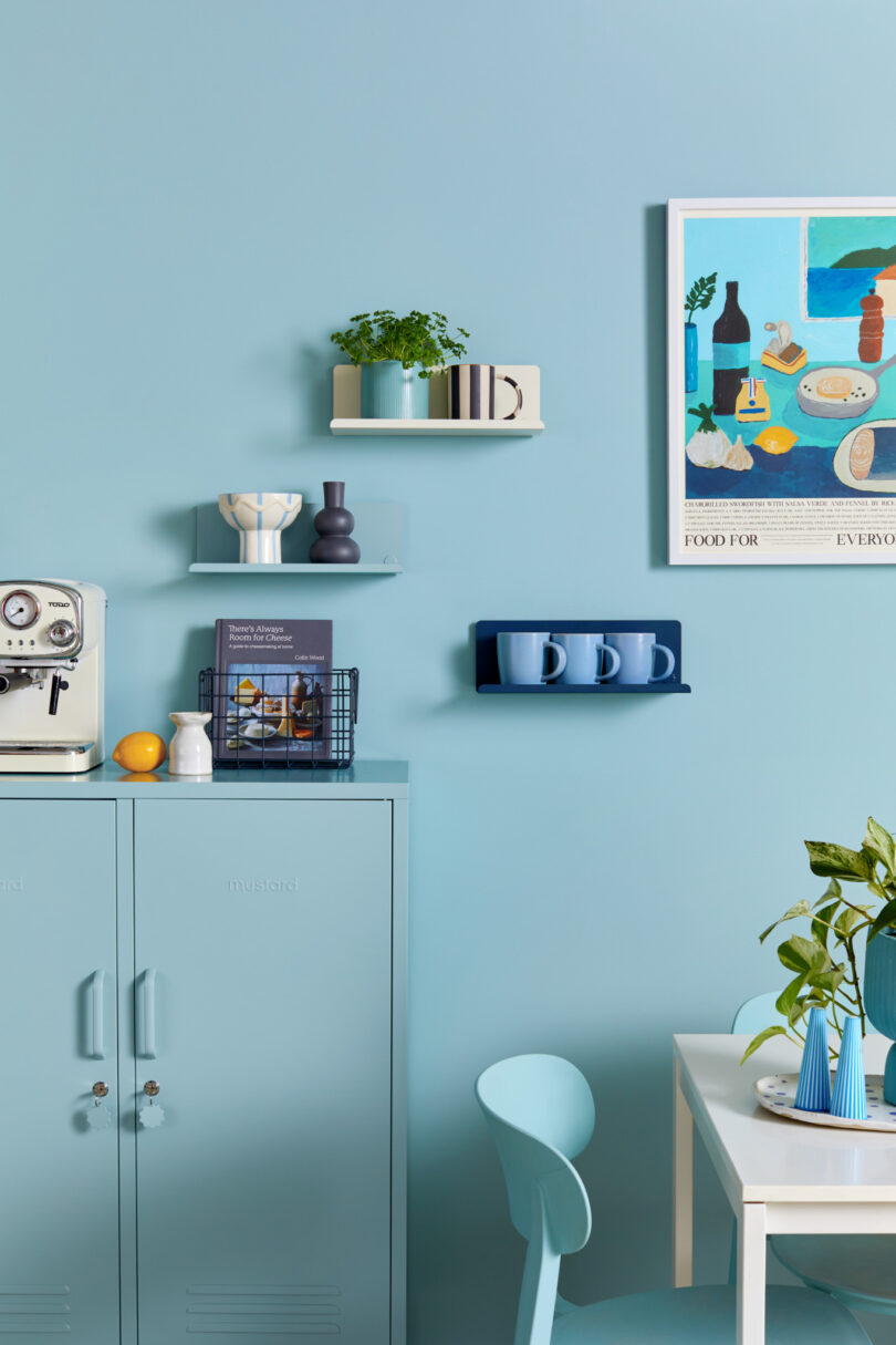 A light blue kitchen with floating shelves holding plants, decor, a small appliance, a book, and mugs. A painting and a cabinet are also present, alongside a table and chair with a plant