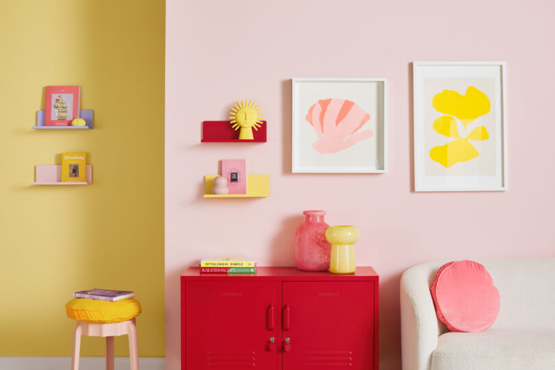 A colorful room with pink and yellow walls, featuring shelves with books and decorations, a red cabinet topped with vases, and framed art above a sofa with a pink cushion