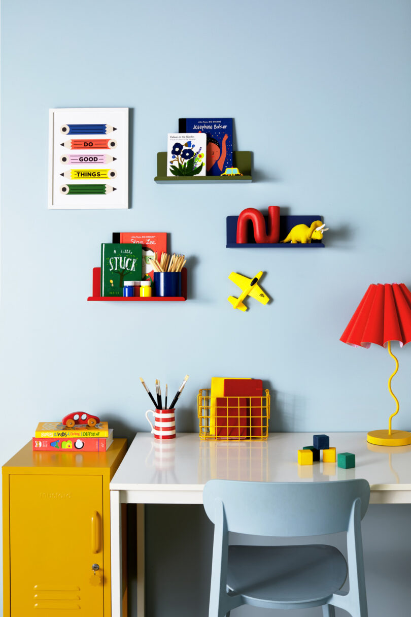 A child's study space with a desk, chair, colorful wall shelves with books, toys, a yellow lamp, and stationery. A yellow cabinet stands beside the desk. The wall is light blue with a framed artwork