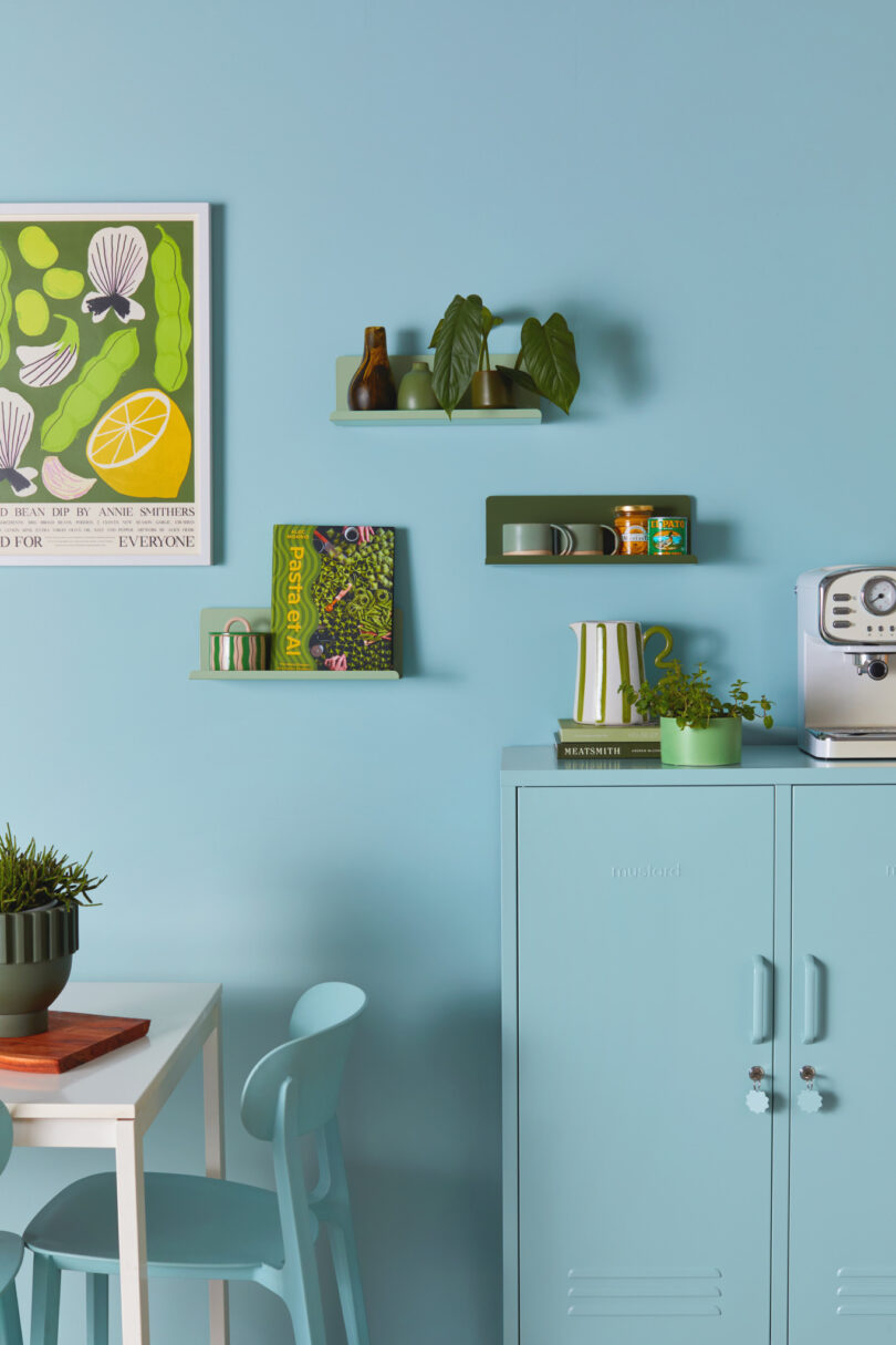 A kitchen with blue walls, a light blue cabinet, green shelves with plants and books, a silver coffee machine, and wall art of fruits