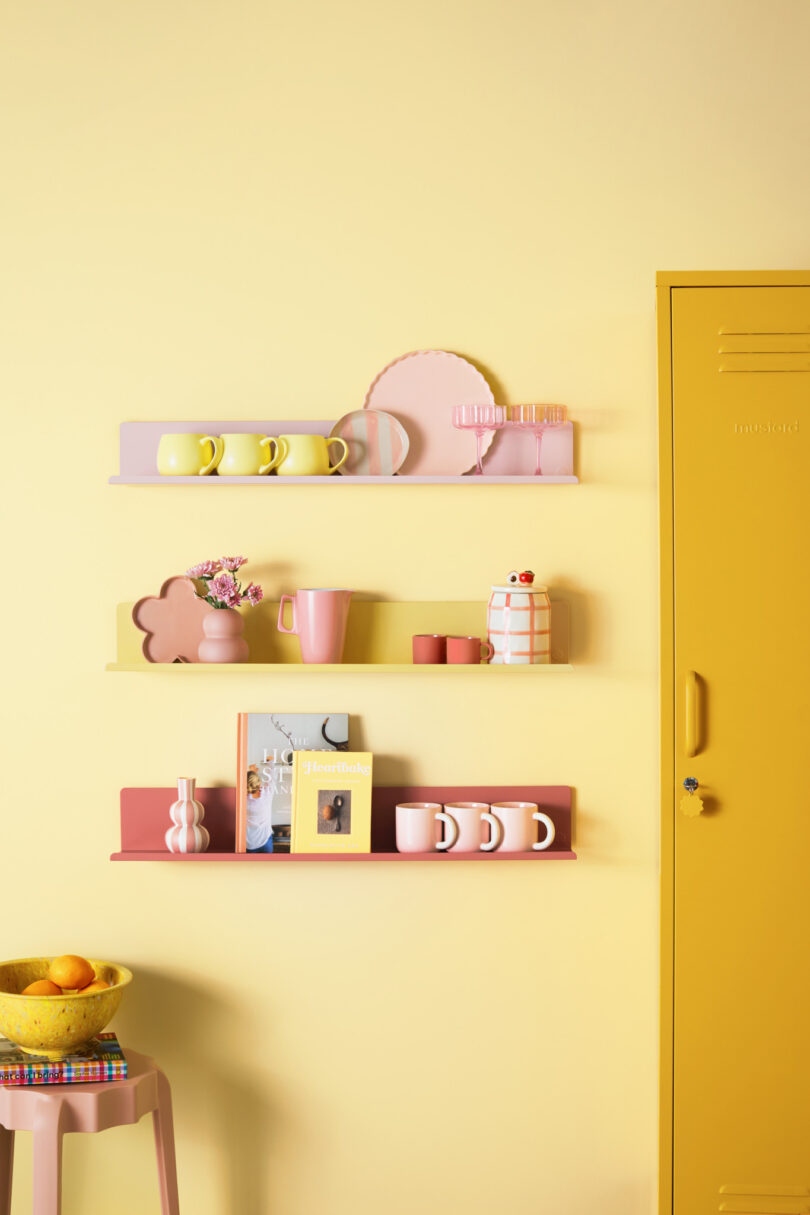 Three colorful wall shelves holding various pastel-colored items, including mugs, a teapot, vases, and books, are mounted on a yellow wall beside a yellow locker. A bowl of fruit is on a small table below