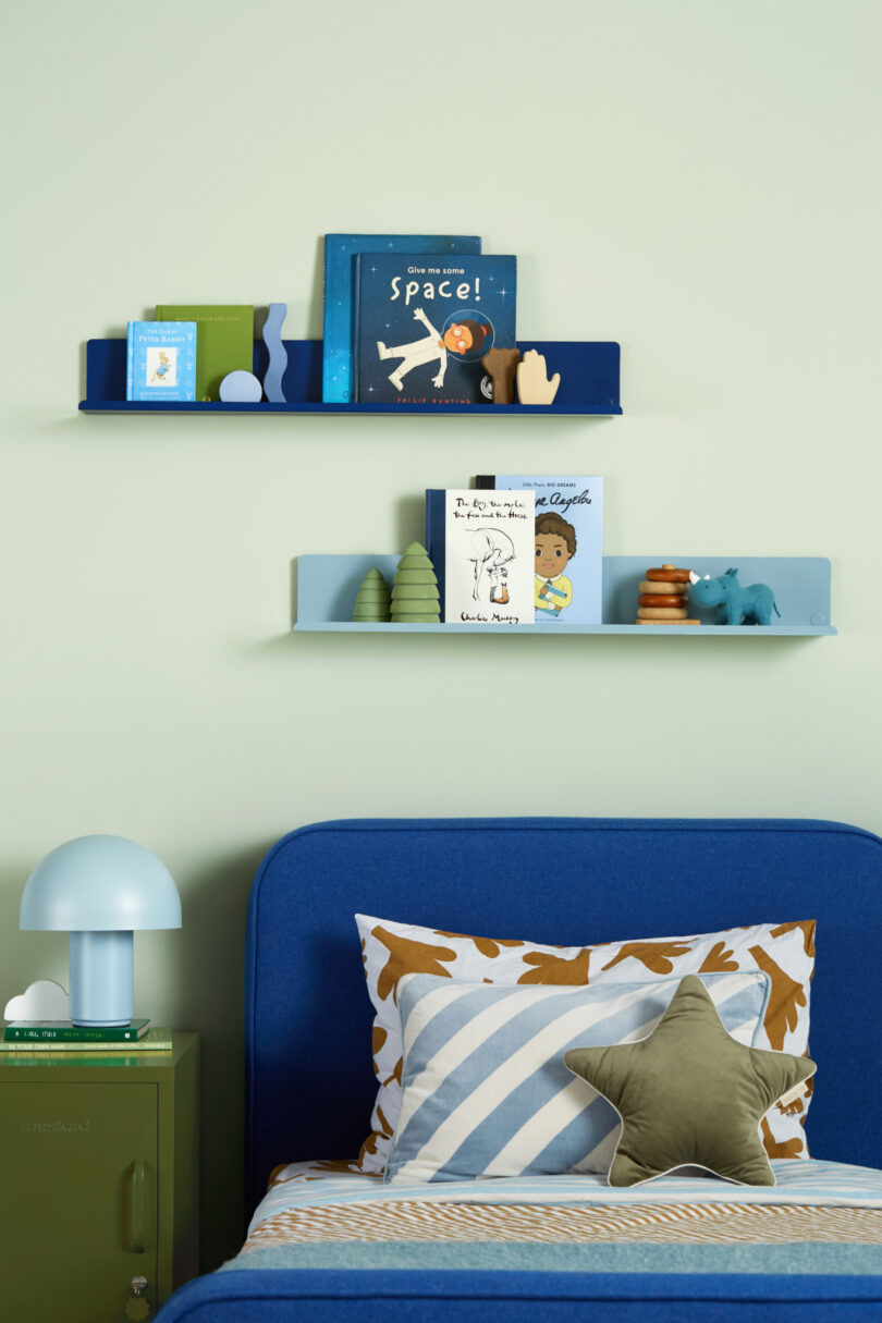 A child's bedroom with shelves holding books and toys above a blue bed with striped bedding, a star-shaped pillow, and a nightstand with a lamp