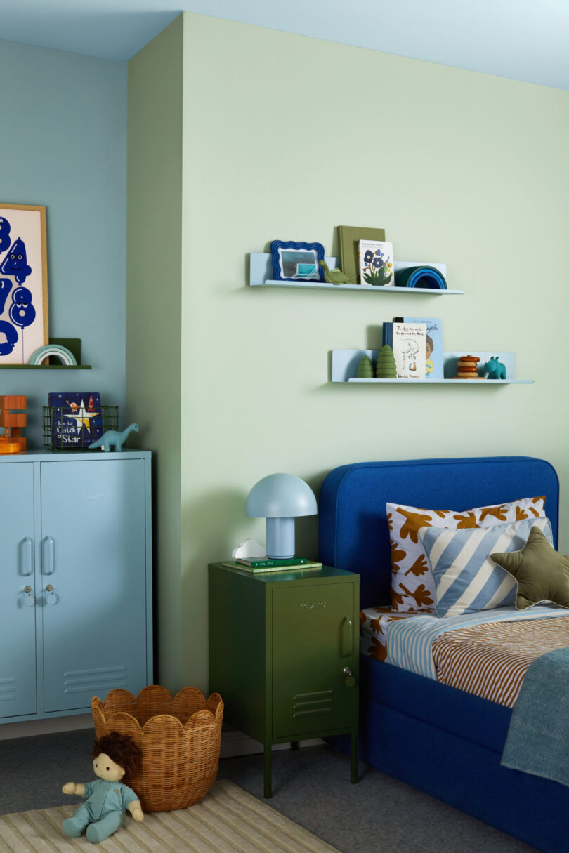 A children's room with a blue bed, blue cabinet, green side table, blue lamp, and shelves with books and toys against light yellow and light blue walls