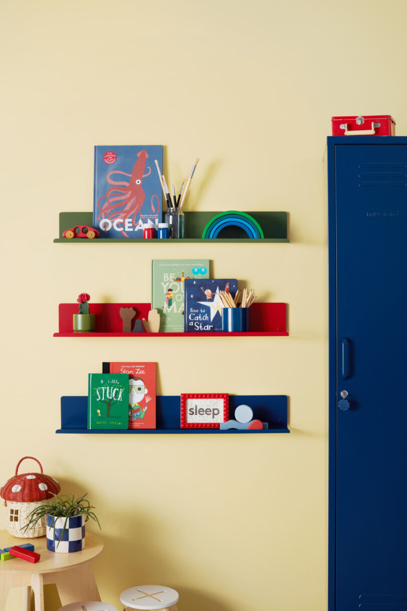 Three colorful wall-mounted shelves displaying children’s books, paint brushes, and toys against a light yellow wall. An adjacent blue locker stands next to them. A small stool is nearby