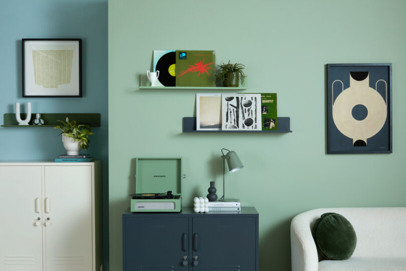 A modern living room with green walls, shelves with records, a plant, and framed art. A record player sits on a dark cabinet, beside a lamp and decor items. A white sofa with a green cushion is in view