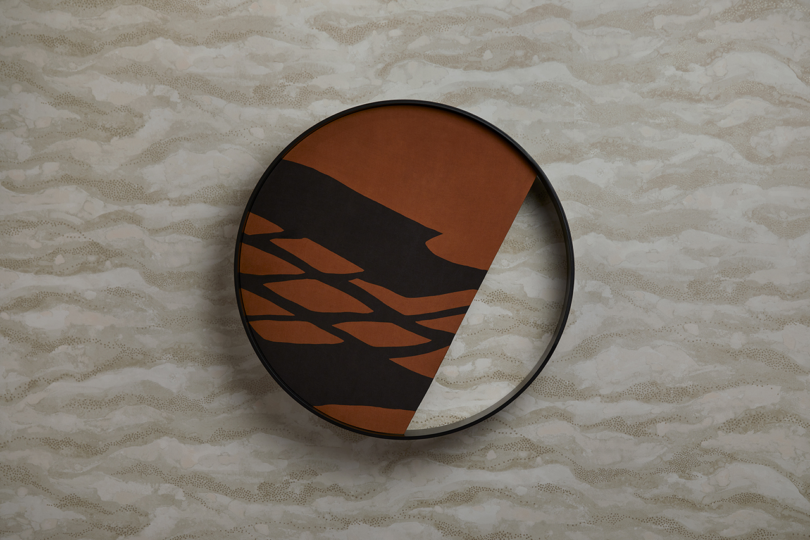 A circular wall art piece with an abstract design in black and brown tones is mounted against a beige patterned wall