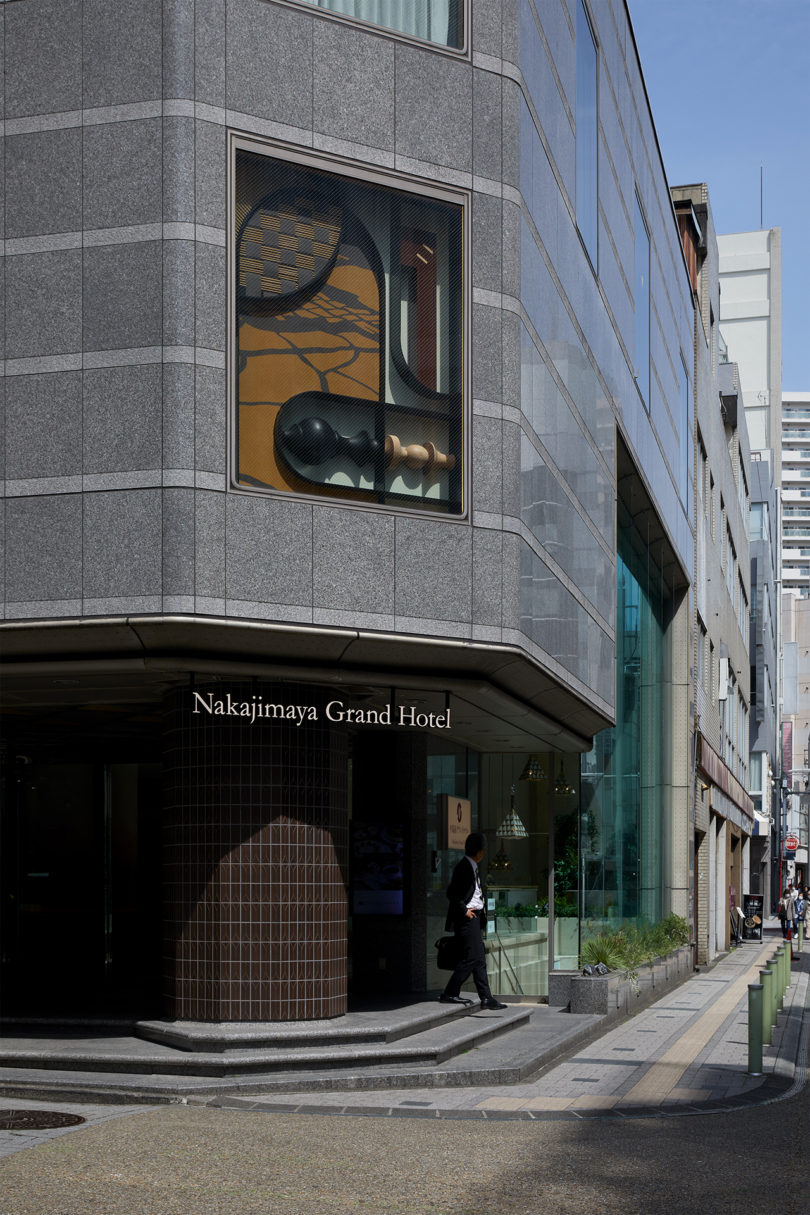 A person walks beneath a corner sign for Nakajimaya Grand Hotel on a modern city street with large windows displaying art pieces