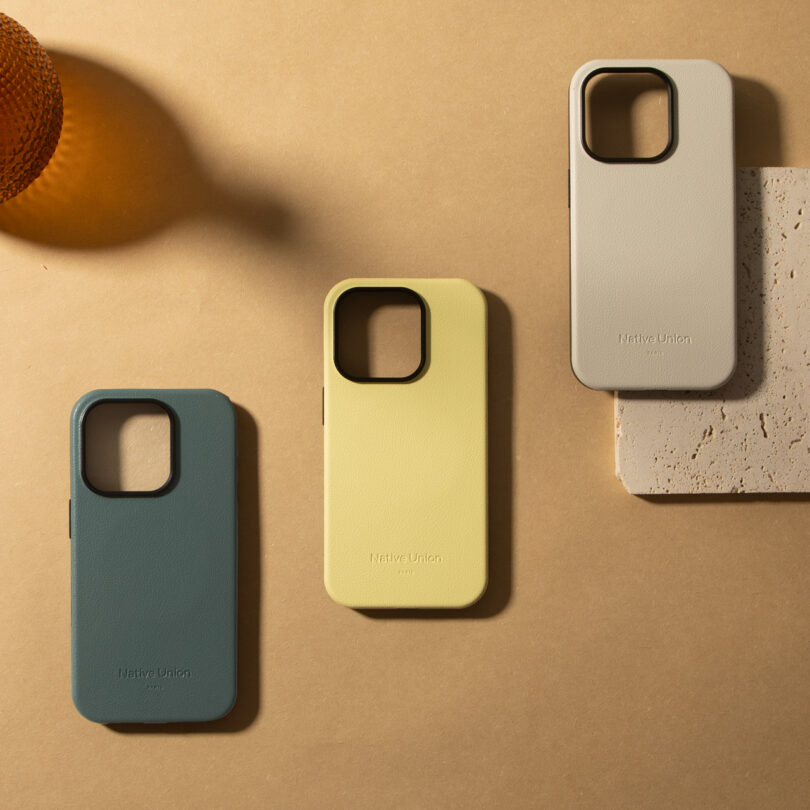 Three smartphone cases in blue, yellow, and beige are displayed on a beige surface next to a textured brown object and a piece of stone.