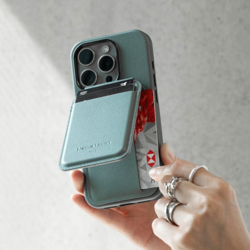 A hand is holding a smartphone with a teal case that has a built-in card holder. A person is pulling a credit card out of the card holder.