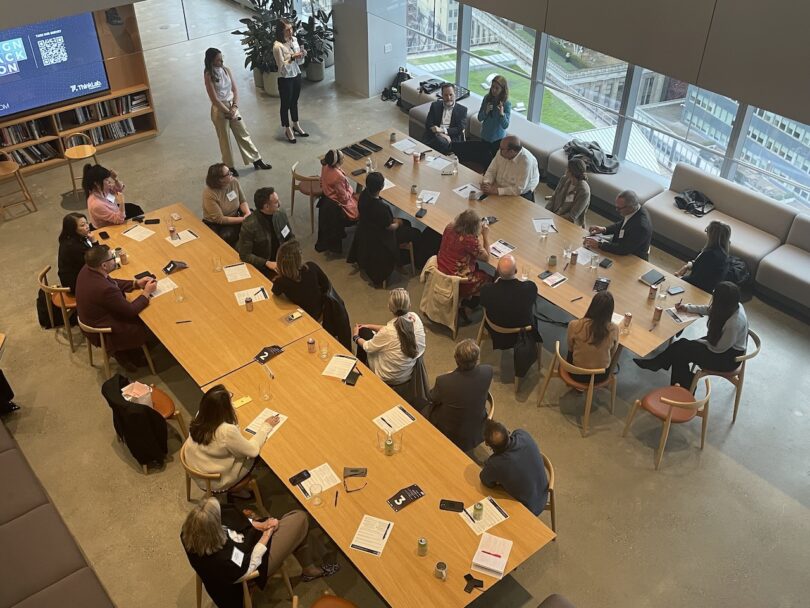 A group of people seated around a large rectangular table in an office setting, engaging in discussion. Some individuals are standing at the front of the room, presenting or speaking to the group.