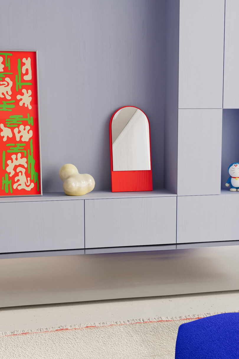 A modern shelf holds an abstract painting, a small sculpture, a red-framed mirror, and a cat figurine. The shelf is part of a minimalist, light gray piece of furniture