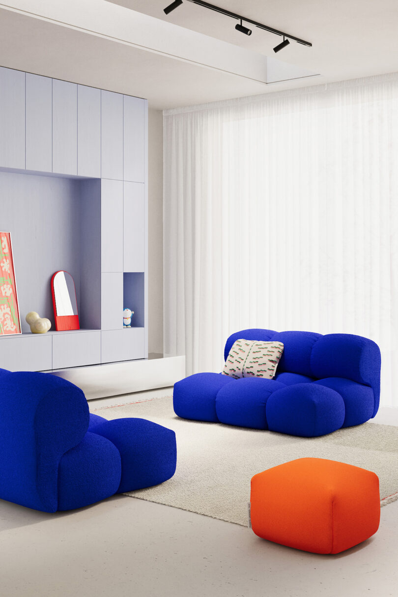 Modern living room with two blue armchairs, an orange ottoman, a beige and white rug, light gray cabinetry, and sheer white curtains. Decorative objects and a mirror are placed on the shelves