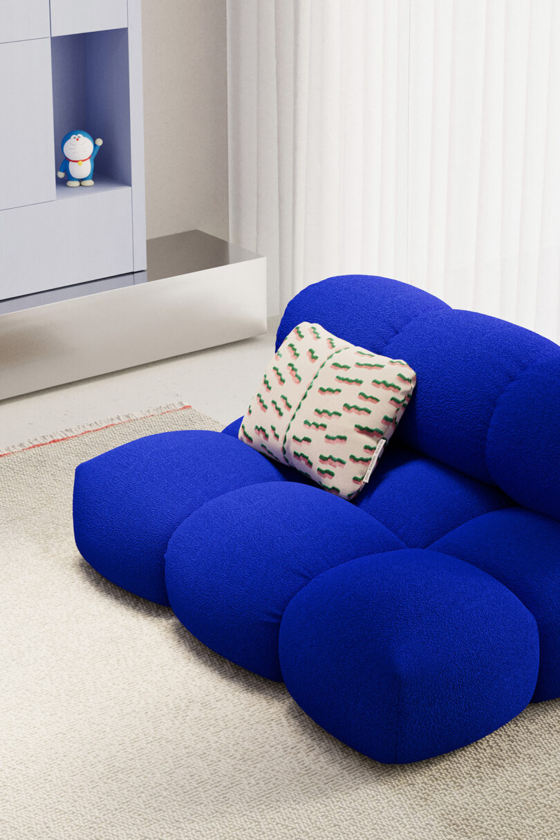 A modern living room with a bright blue plush couch and a patterned pillow. A cabinet on the left displays a small toy. White curtains and a light-colored carpet are also visible