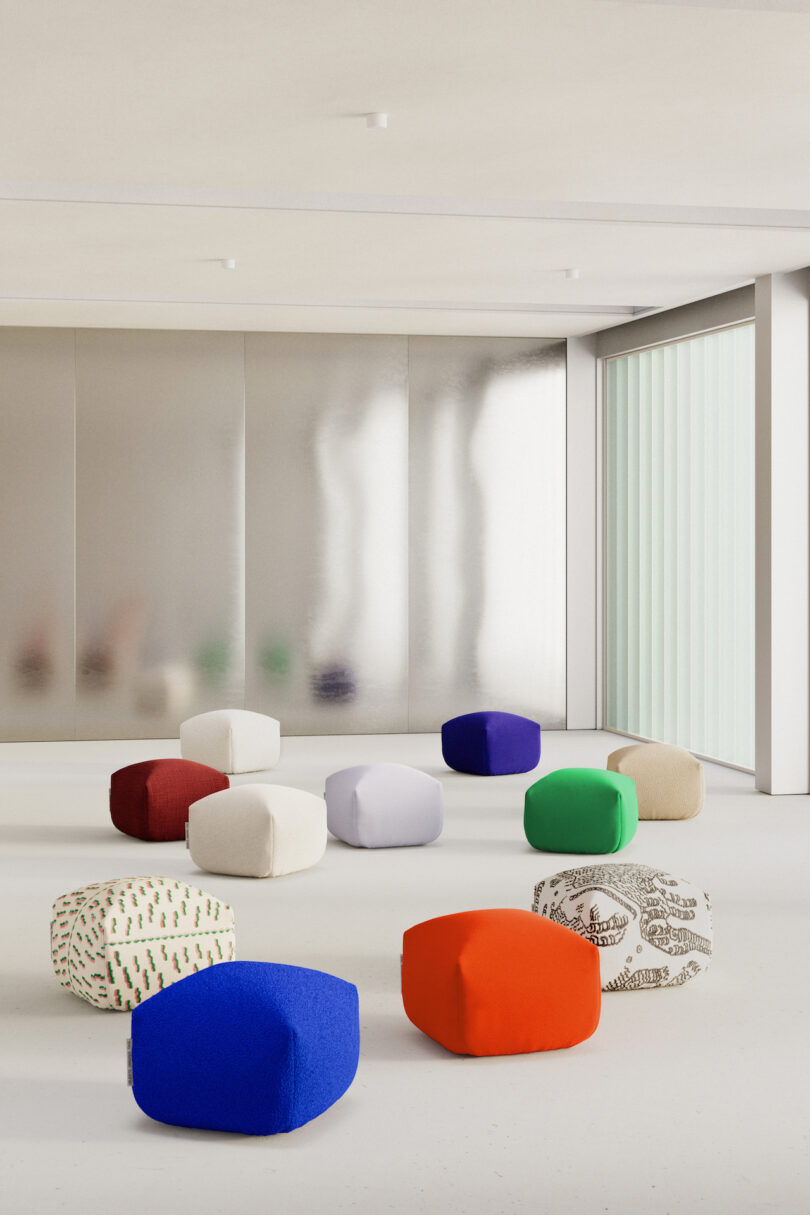 A minimalist room features multiple colorful, geometric poufs arranged on the floor. The background includes a large frosted glass panel and a window with vertical blinds.