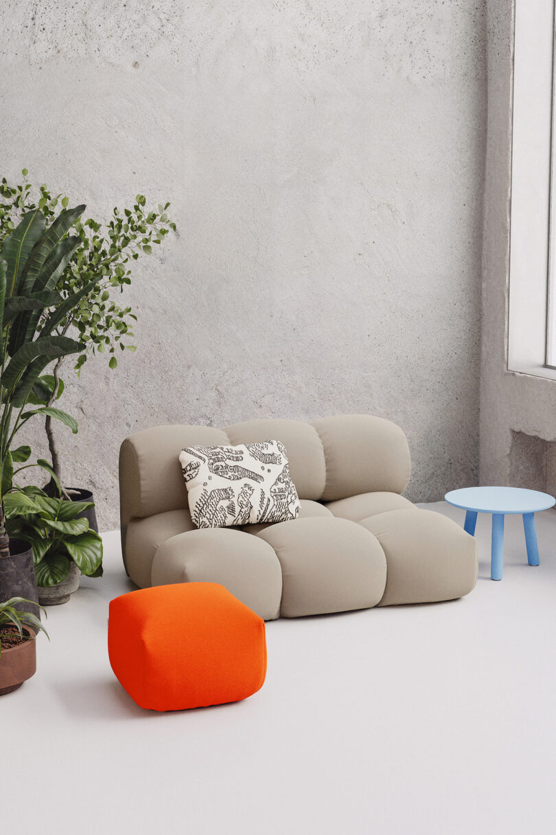 A modern seating arrangement featuring a beige modular sofa, an orange ottoman, a blue side table, and potted plants near a large window with natural light