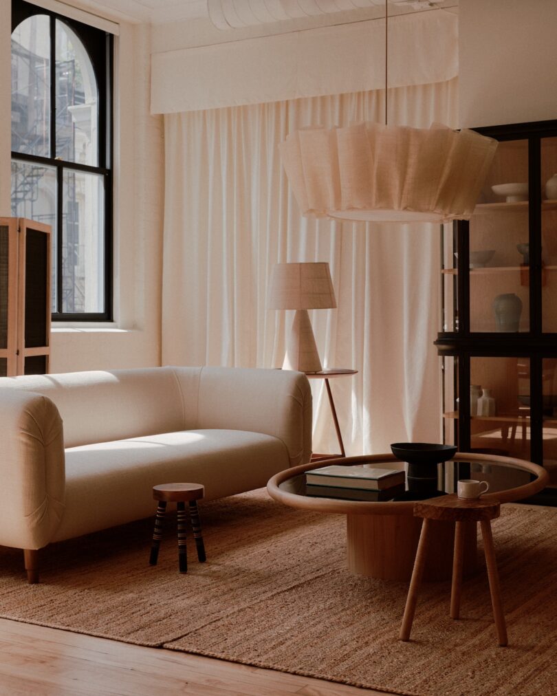 A minimalist living room with a white sofa, round coffee table, stool, and a large pendant light. Neutral colors dominate the space, featuring a window with sheer curtains and a woven rug.