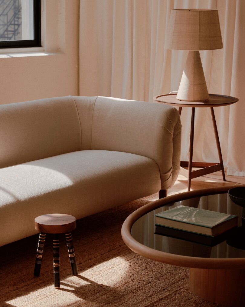 A modern living room features a light beige sofa, a round coffee table with books, a small wooden stool, and a lamp on a side table. Natural light streams in through a window with sheer curtains.