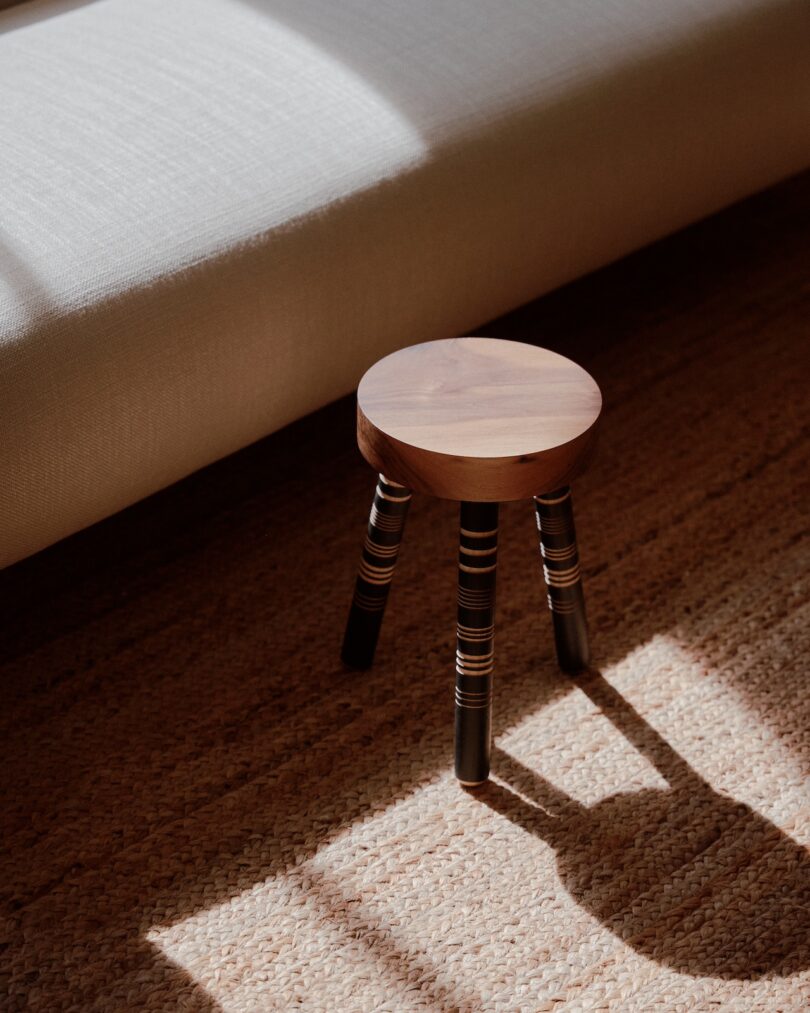 Small wooden stool with striped legs placed on a woven rug near a white sofa, illuminated by sunlight.