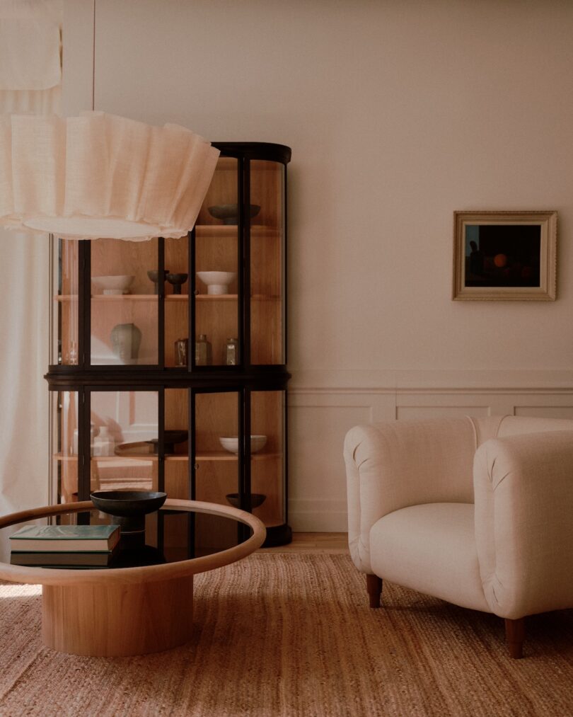 A cozy living room features a white armchair, a round coffee table with books and bowls, and a wooden cabinet with glass doors. A framed painting hangs on the wall above the chair.