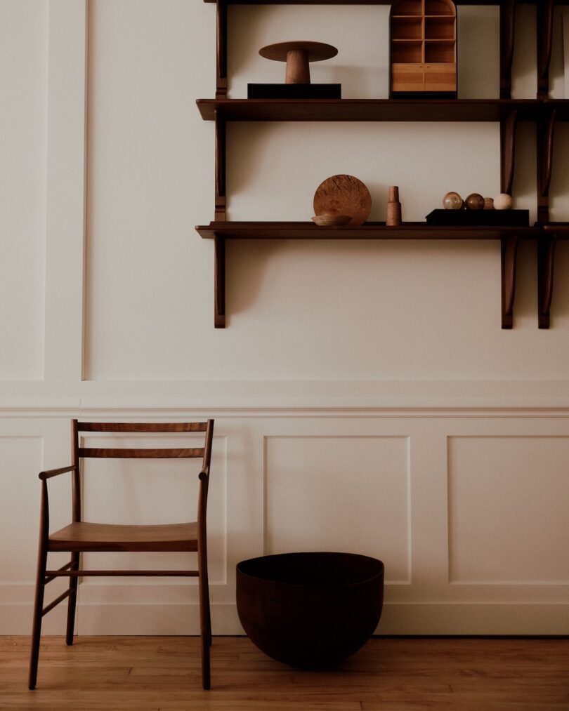 Minimalist interior with wooden chair and shelves adorned with decorative items; large bowl on the floor.