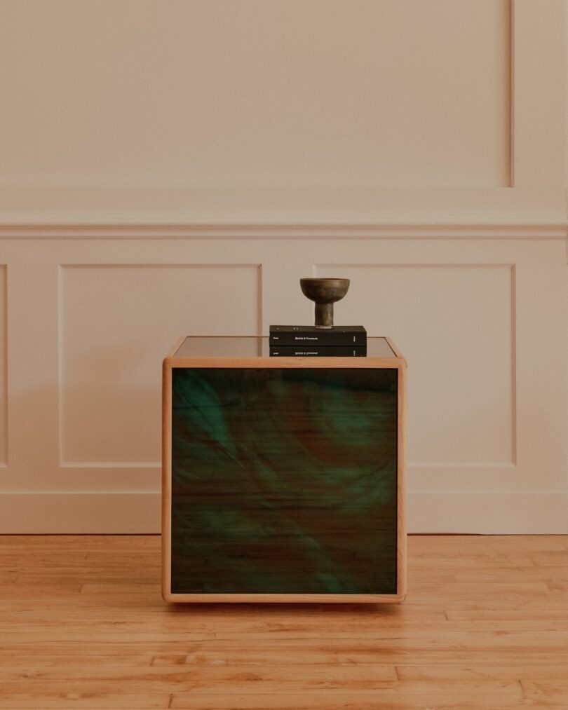 A wooden side table with green-tinted drawer fronts, two black books, and a small decorative object on top, placed against a cream-colored paneled wall on a hardwood floor.