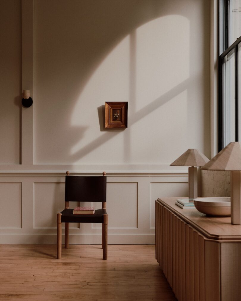 An empty chair sits in front of a paneled wall with a small framed picture. A side table with lamps and decorative items is nearby, illuminated by natural light from a large window.