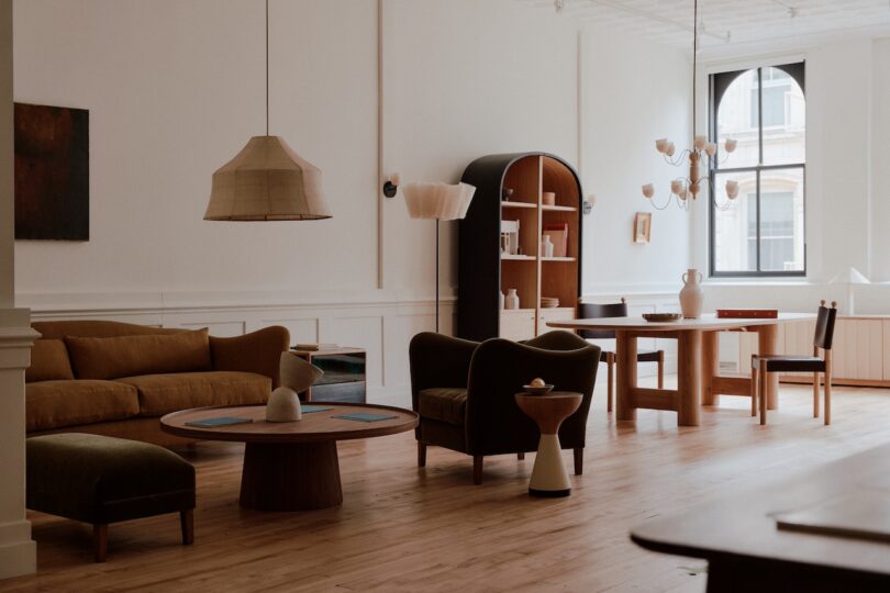 A modern living room with light wooden floors, beige and green furniture, a round dining table, bookshelves, a large window, and minimalistic decor.