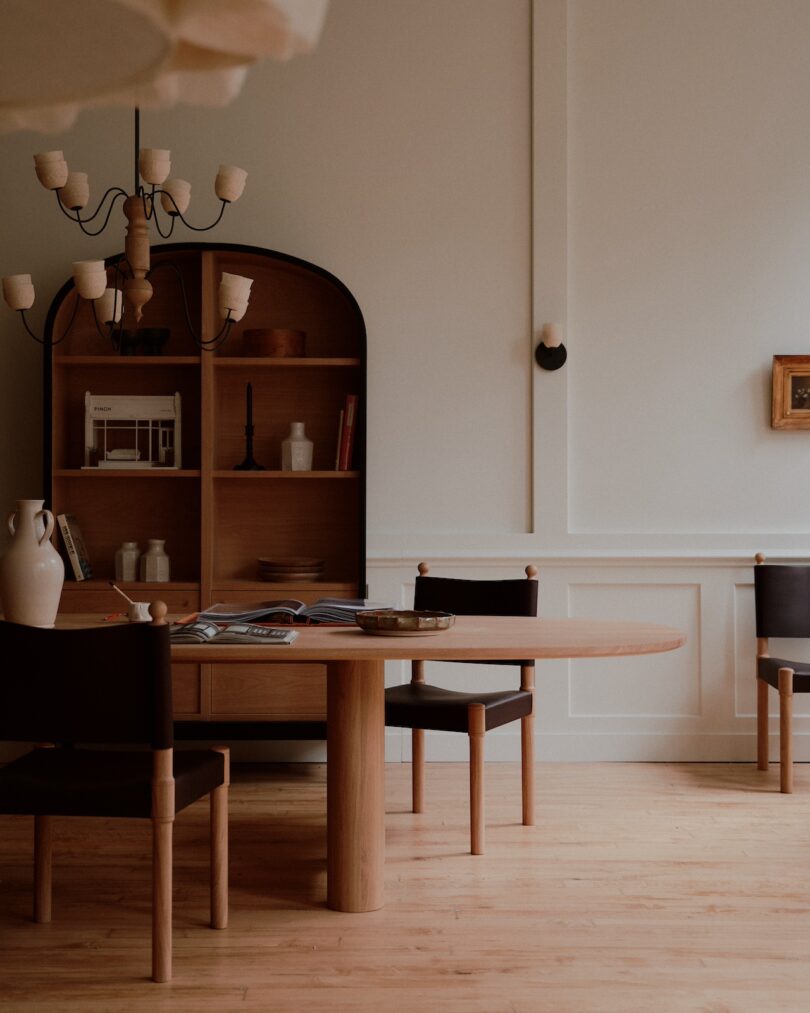 A minimalist dining room features a wooden table with a book and ceramics, black chairs, a chandelier, a shelf with books and decor, and a small wall-mounted picture.