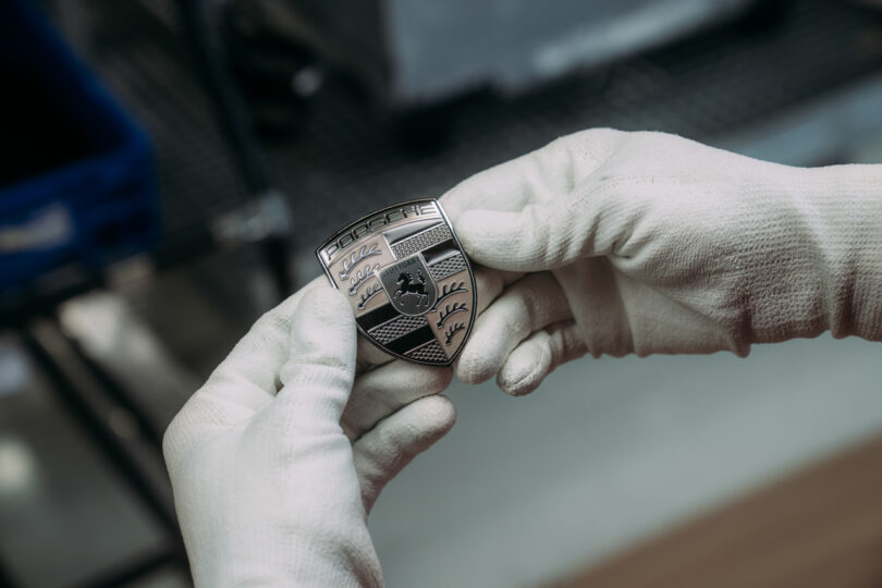 Close-up of gloved hands holding a metallic Porsche emblem with a black and silver design.