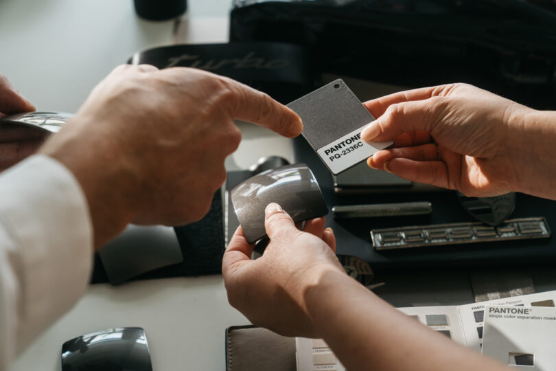 Two people discuss and compare Pantone color samples, focusing on a grey shade labeled "PANTONE PQ-2390C." Various color samples and design materials are seen on the table.