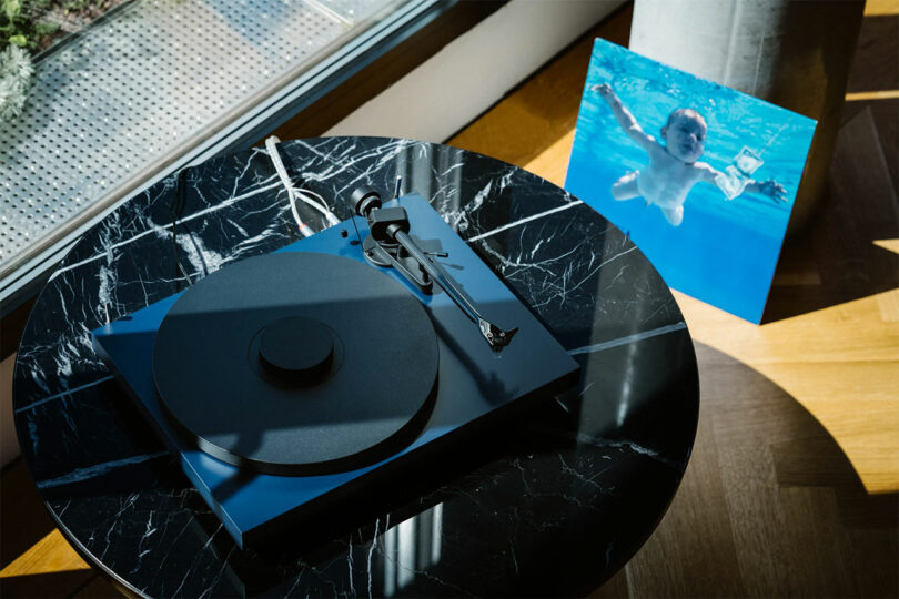 A black Pro-Ject Debut EVO 2 turntable sits on a round marble table, next to a vinyl record cover depicting a baby swimming underwater.