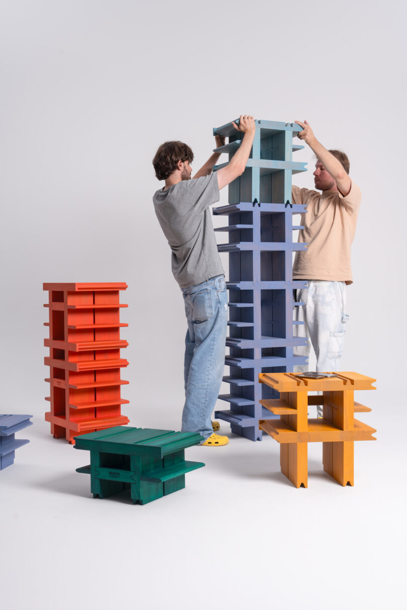 Two people are assembling a tall, blue modular shelving unit. Surrounding them are other colorful, disassembled shelving units in red, green, and yellow