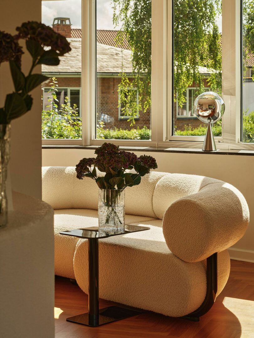 A cozy living room with a curved white sofa, a glass vase with dark flowers on a black side table, and large windows showing an outdoor view of greenery and a brick building