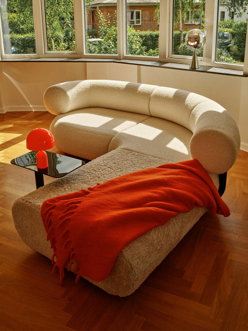 A plush, white curved sofa with an attached ottoman topped with a red blanket next to a small black side table holding an orange lamp in a sunlit room with a large window and wooden floor