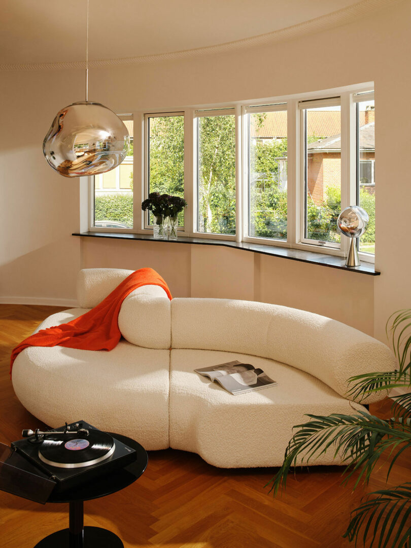 A modern living room features a curved white sofa with an orange throw blanket, a coffee table with a vinyl record player, and a large window letting in natural light