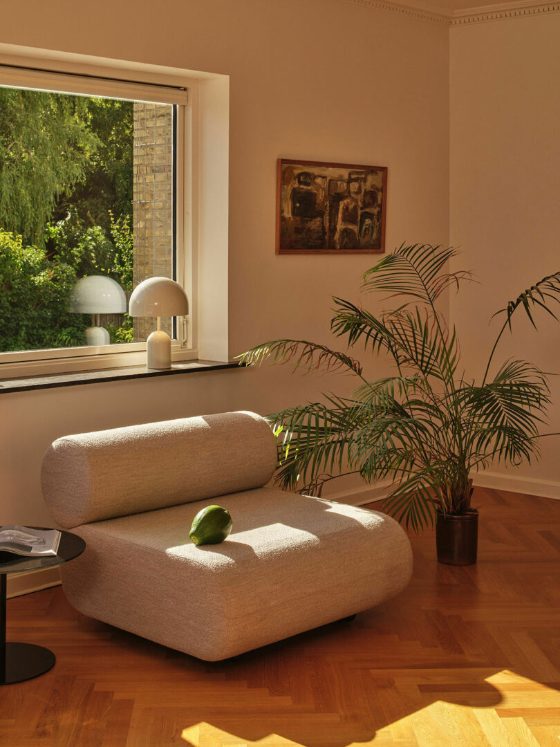 A modern living room with a light gray chair, a small side table, a large potted plant, and a window with two table lamps. Sunlight illuminates the room, highlighting the green plant and wooden floor