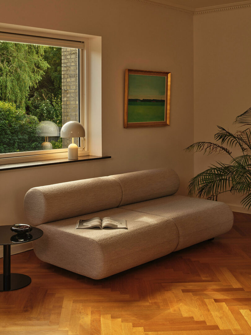 A minimalist living room with a gray sofa, an open book, a round black side table, a large window showing greenery, and a framed picture on the wall