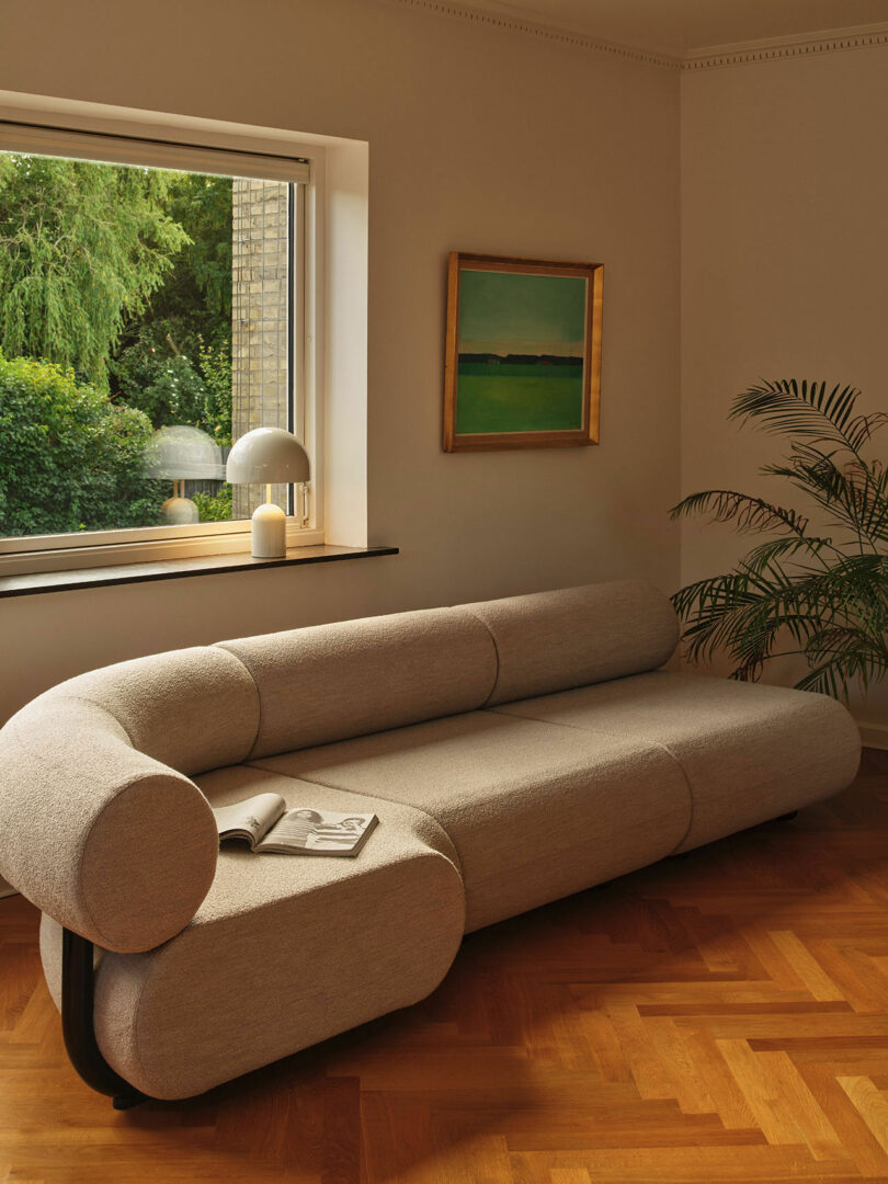 A beige sofa with a magazine on it is placed in a warmly lit living room with a large window, a green landscape painting, and a houseplant