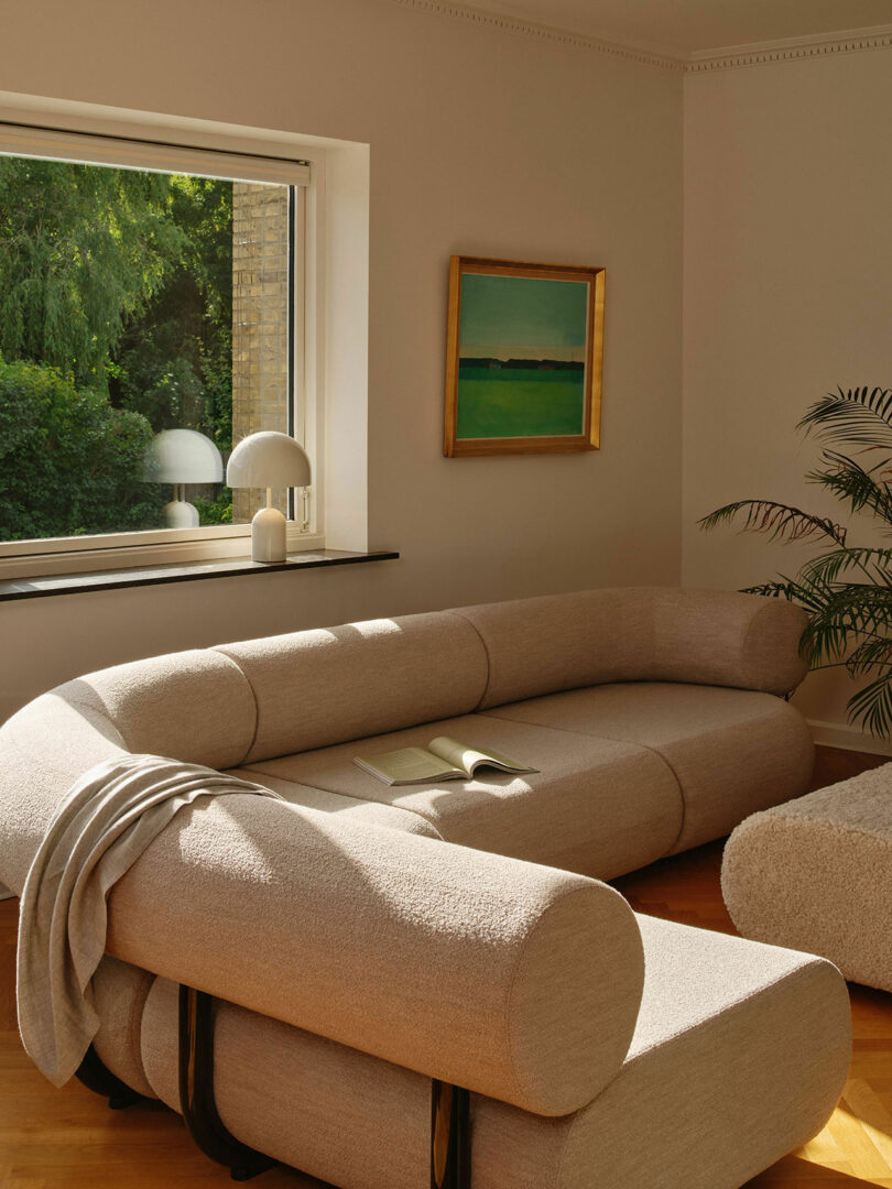 A minimalist living room with a beige sectional sofa, a small table lamp, an open book, a window showing greenery, a framed painting on the wall, and a potted plant in the corner