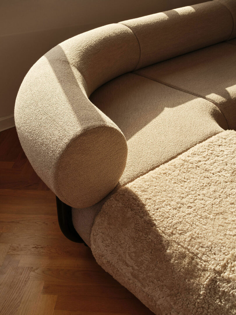 A close-up of a beige, modern sofa with a textured fabric finish and a round armrest, illuminated by natural sunlight on a wooden floor