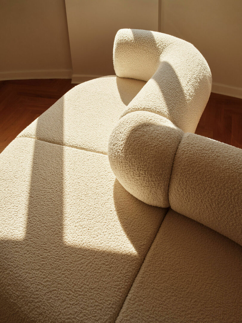 A beige, boucle curved sofa is illuminated by sunlight, casting shadows on its textured surface. The sofa is set against a wooden floor and a light-colored wall