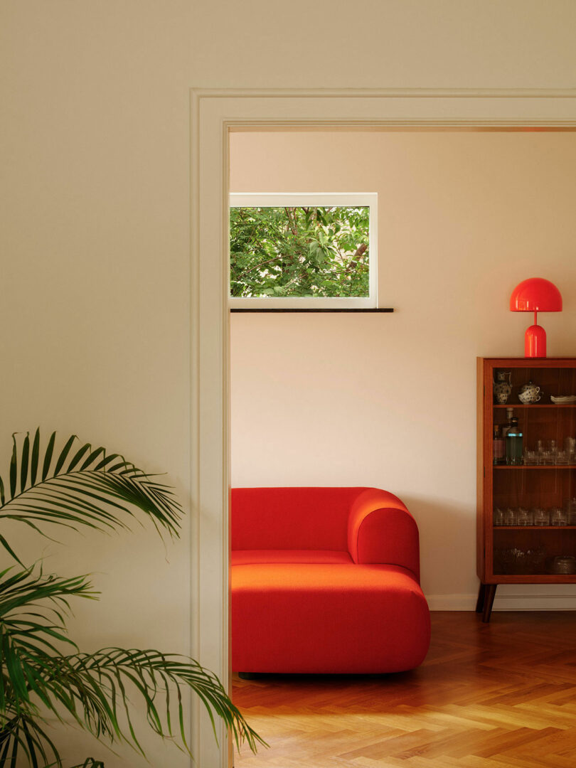 A cozy living room with a bold red sofa, a wooden cabinet with glassware, a red lamp, and a large leafy plant, viewed from an adjacent room's doorway. A small window shows greenery outside