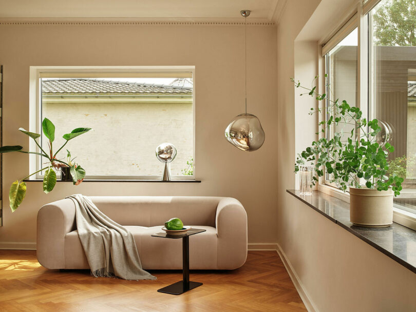 A modern living room with a beige couch, a side table holding a green apple, large windows, and potted plants on the window sills. Natural light illuminates the space