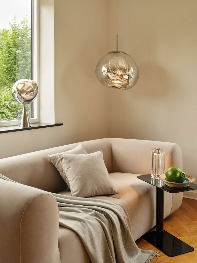 A modern living room features a beige sofa with a throw blanket and cushion, a black side table holding a glass of water and green fruit, a metallic desk lamp, and a pendant light near a window